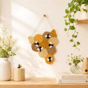 May include: A wall hanging featuring a cluster of crocheted hexagonal honeycomb shapes in shades of yellow, gold, and tan. Four small, crocheted bees with black and yellow striped bodies and white wings are attached to the honeycomb structure, which is suspended by a thin string.