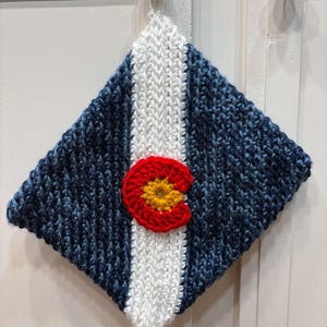 May include: A diamond-shaped, hand-knitted potholder featuring a vertical white stripe centered between two dark blue sections. A circular, crocheted emblem in red and yellow is attached to the center of the white stripe, creating a distinct design.