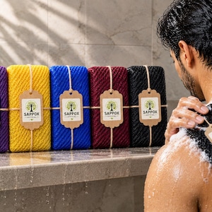 Puede incluir: Una colección de esponjas de baño rectangulares y texturizadas en morado, amarillo, azul, granate y negro. Cada esponja tiene una etiqueta de papel marrón atada con cordel, con un logotipo de árbol y el texto SAPPOX OFFICIAL.