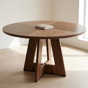 May include: A round, dark brown wooden dining table with a smooth, circular top and a sturdy, geometric base featuring four angled legs that meet in a central cross-brace. A small, rectangular, light-coloured book rests flat on the centre of the table.