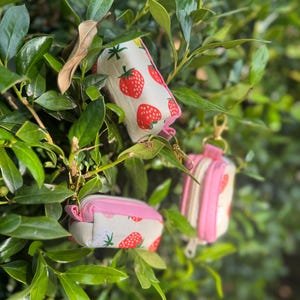 May include: Three small, rectangular pouches made of off-white fabric featuring a red strawberry pattern with green leaves. Each pouch has a bright pink zipper closure and a gold-toned metal lobster clasp attached to a pink fabric loop.
