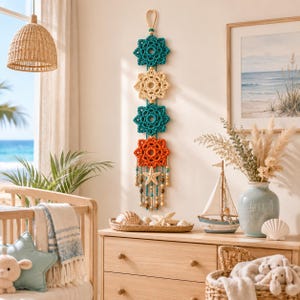 May include: A vertical wall hanging featuring four crocheted floral motifs in teal, cream, and orange, suspended from a loop. The piece includes dangling strands adorned with wooden beads and a small starfish at the bottom center.