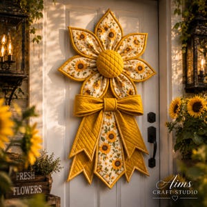 May include: A quilted fabric sunflower wall hanging featuring a central yellow textured disc, petals with a sunflower print on a cream background, and a large mustard yellow bow with long, pointed, quilted fabric tails hanging downwards.