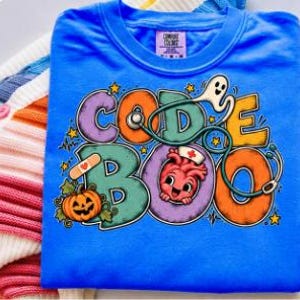 Puede incluir: Una camiseta azul brillante con un gráfico colorido y el texto CODE BOO. El diseño incluye un estetoscopio, un fantasma, una calabaza, una tirita y un corazón de dibujos animados con una cofia de enfermero. Etiqueta Comfort Colors.