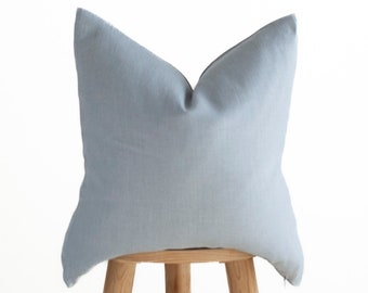 May include: A light blue square pillow sits on a wooden stool. The pillow is made of a soft, textured fabric.