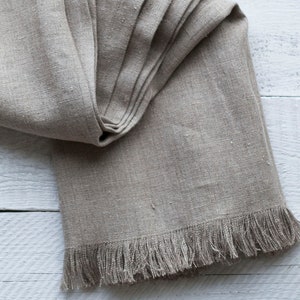 May include: A folded linen table runner with a fringed edge, displayed on a white wooden surface.