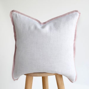 Peut inclure: Un oreiller en lin blanc avec une bordure rose. L'oreiller est posé sur un tabouret en bois.