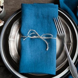 May include: A blue linen napkin tied with twine, placed on a silver and black plate with a fork. The plate is on a wooden table with a blue linen tablecloth and dried flowers.