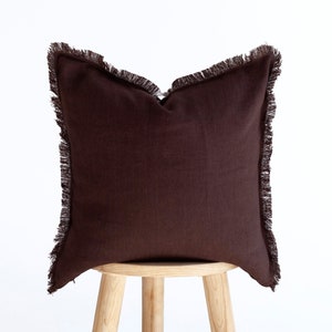 May include: A brown square pillow with a fringe border. The pillow is sitting on a wooden stool.
