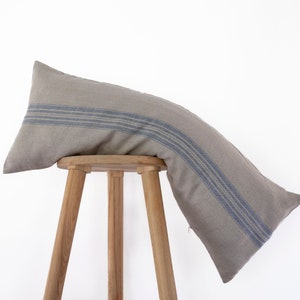 May include: A long, gray linen pillow with blue stripes sits on a wooden stool. The pillow is slightly curved and has a relaxed, casual look.