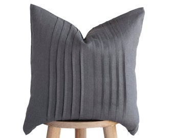 May include: A grey pleated cushion sits on a wooden stool. The cushion is made of a soft, textured fabric and has a simple, elegant design.