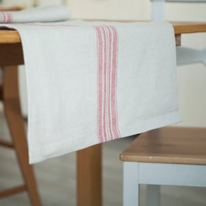 Peut inclure: Un chemin de table en lin blanc avec trois bandes rouges au centre. Le chemin de table est posé sur une table en bois.