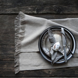 May include: A rustic table setting with a grey linen placemat, a silver plate, a black and white plate, a silver knife and fork, and a grey napkin tied around a white egg with a floral design.