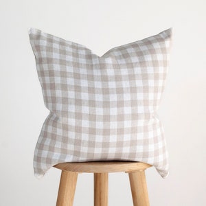 May include: A beige and white gingham pillow sits on a wooden stool. The pillow is made of a soft, textured fabric and has a square pattern.