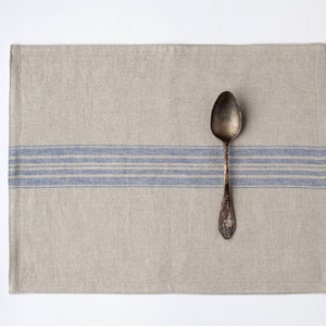 May include: A beige linen placemat with a blue stripe design and a vintage silver spoon. The placemat is rectangular and made of textured fabric. The spoon is placed on the right side. Table setting.