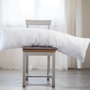 May include: A white body pillow resting on a grey chair with a light grey cushion. The chair is in a room with white curtains and a white wall.