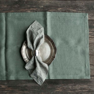 May include: A sage green linen table runner with a silver plate and a linen napkin with a silver napkin ring. The table runner is on a rustic wooden surface.