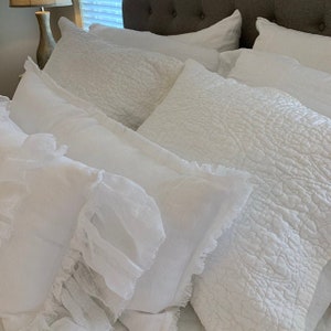 May include: A collection of white, ruffled pillowcases with a textured, quilted design. The pillowcases are arranged on a bed with a brown headboard.
