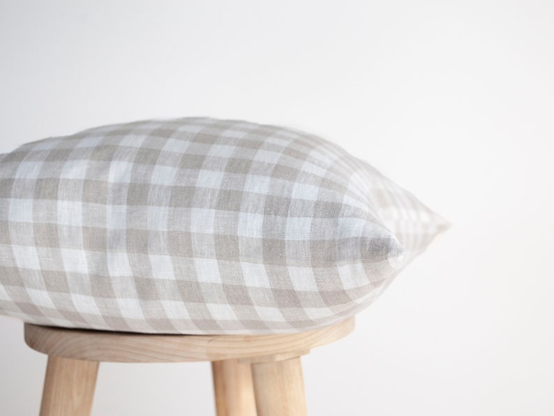 May include: A beige and white gingham pillow resting on a wooden stool. The pillow is made of a soft, textured fabric.
