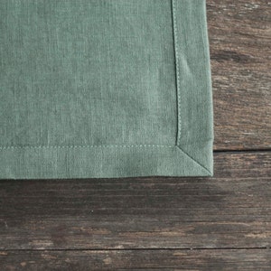May include: A close-up of a green linen napkin with a stitched edge. The napkin is lying on a wooden surface.