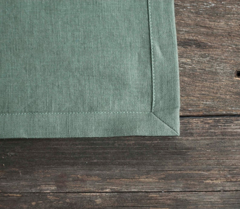 May include: A close-up of a green linen napkin with a stitched edge. The napkin is lying on a wooden surface.