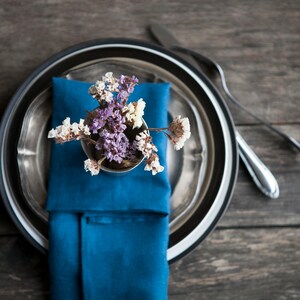 Peut inclure: Une serviette bleue avec des fleurs s&eacute;ch&eacute;es dans un petit vase argent&eacute; repose sur une assiette argent&eacute;e et noire. L'assiette est sur une table en bois.