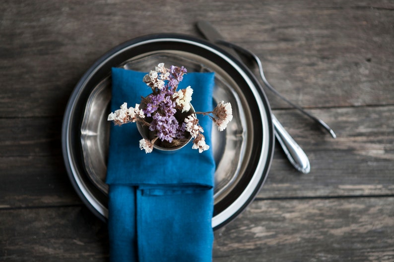 Peut inclure: Une serviette bleue avec des fleurs s&eacute;ch&eacute;es dans un petit vase argent&eacute; repose sur une assiette argent&eacute;e et noire. L'assiette est sur une table en bois.