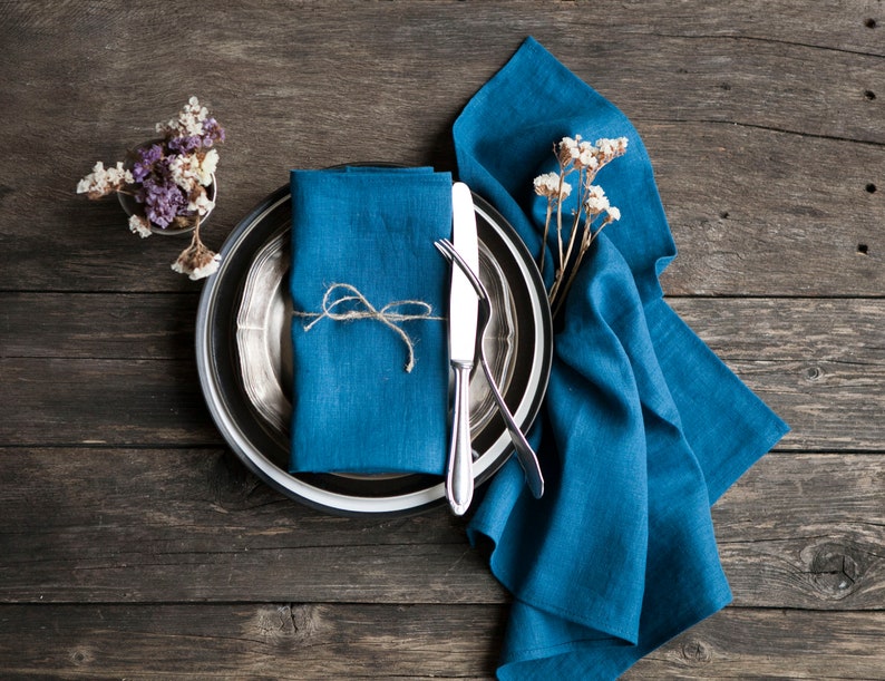 Peut inclure: Une serviette en lin bleu nou&eacute;e avec de la ficelle, plac&eacute;e sur un chargeur argent&eacute; avec un bord noir, et un couteau et une fourchette en argent. Le d&eacute;cor est sur une table en bois rustique.