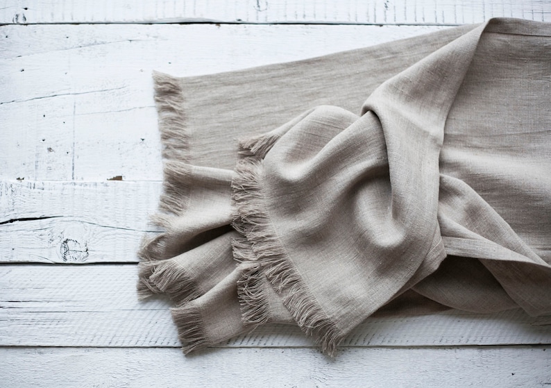 May include: A beige linen throw blanket with fringed edges, folded and draped on a white wooden surface.