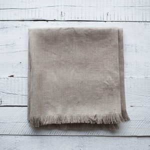 May include: A beige linen table runner with a fringed edge, laid flat on a white wooden surface.