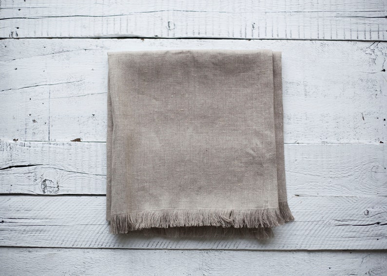 May include: A beige linen table runner with a fringed edge, laid flat on a white wooden surface.