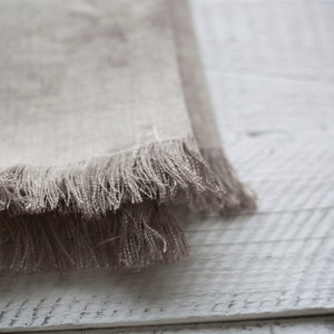May include: A close-up of a light brown linen fabric with a frayed edge. The fabric is folded over itself and has a soft, textured appearance.