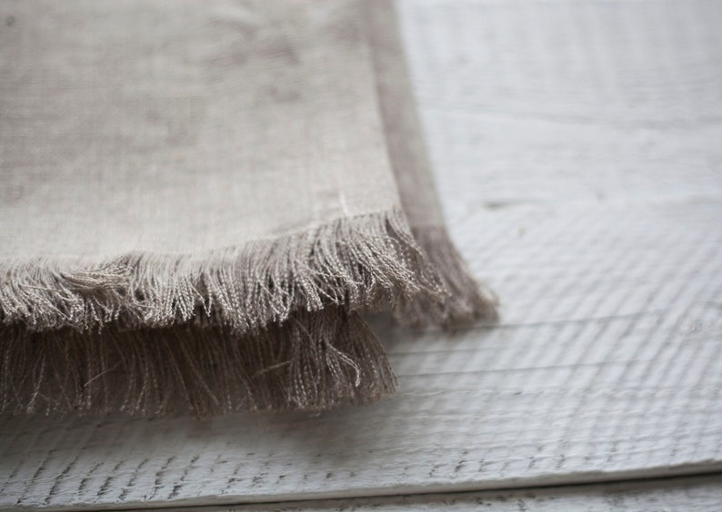 May include: A close-up of a light brown linen fabric with a frayed edge. The fabric is folded over itself and has a soft, textured appearance.
