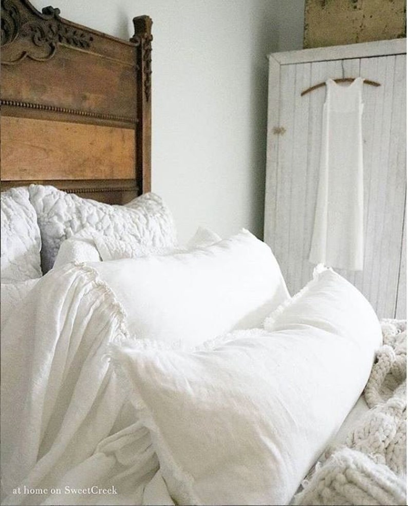 May include: A white bed with a wooden headboard and white bedding. The bed is made with a white duvet cover and several white pillows. A white bolster pillow is on the bed. The bed is in a bedroom with a white closet in the background.