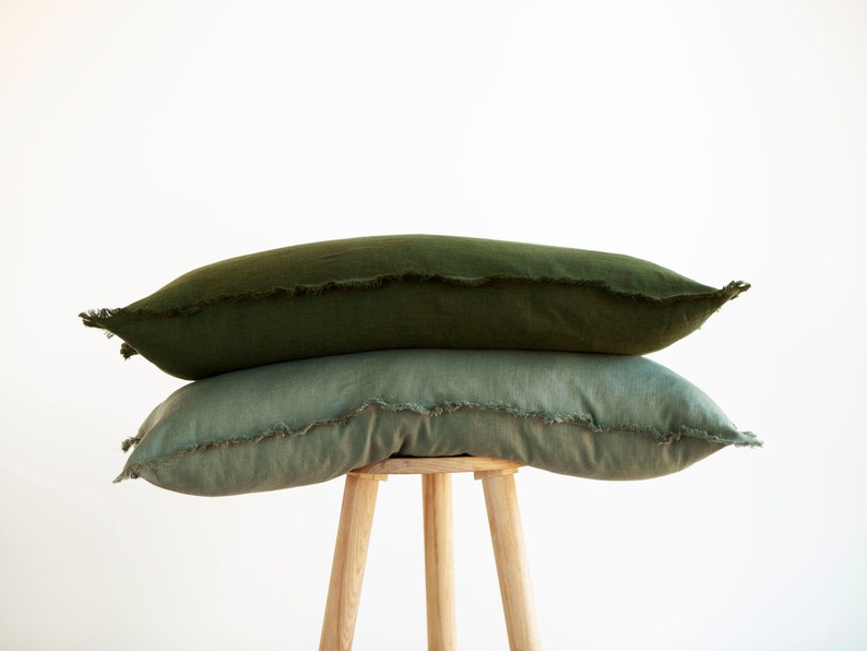 May include: Two green linen throw pillows with frayed edges stacked on a wooden stool.