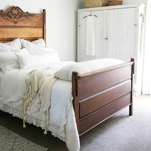 May include: A white bedroom with a wooden bed with a white bedspread and a white blanket. The bed has a carved wooden headboard. There is a white closet with a white door and a white dress hanging on a hanger inside. There is a rug on the floor.