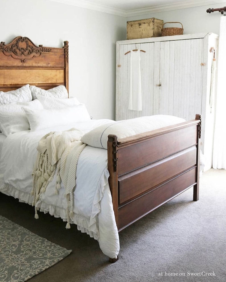 May include: A white bedroom with a wooden bed with a white bedspread and a white blanket. The bed has a carved wooden headboard. There is a white closet with a white door and a white dress hanging on a hanger inside. There is a rug on the floor.