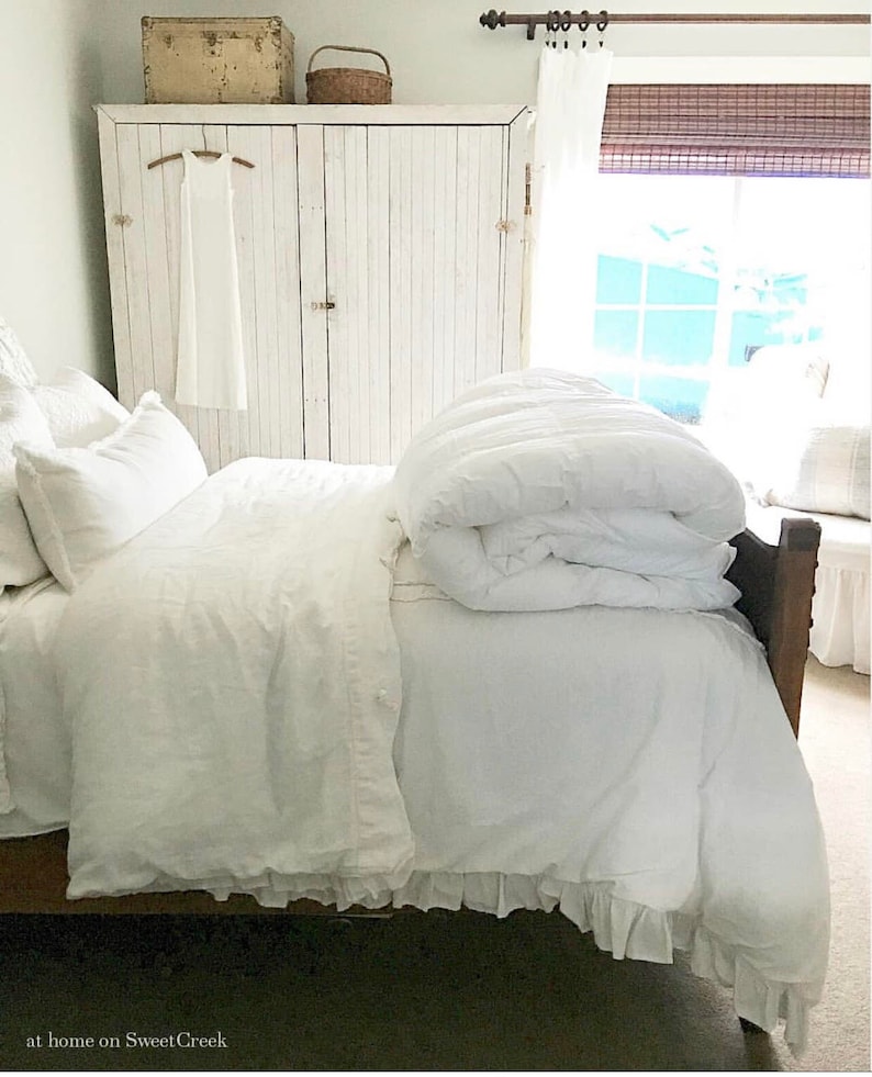 May include: A white bed with a white duvet and a white ruffled bed skirt. The bed is in a bedroom with white painted cabinets and a window with a brown bamboo shade.