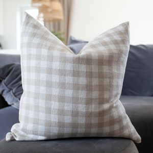 May include: A beige and white gingham throw pillow with a soft, textured fabric. The pillow is sitting on a gray couch.