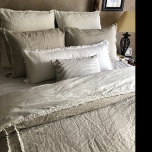 May include: A bed with a white linen duvet cover and a variety of pillows in shades of white and beige. The pillows have a frayed edge detail and the duvet cover has a subtle texture.