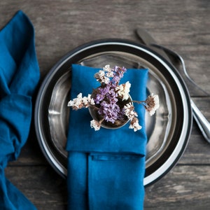 Peut inclure: Une serviette bleue est pli&eacute;e et plac&eacute;e sur une assiette en argent avec un bord noir. Un petit bouquet de fleurs s&eacute;ch&eacute;es est plac&eacute; sur la serviette. L'assiette est sur une table en bois.