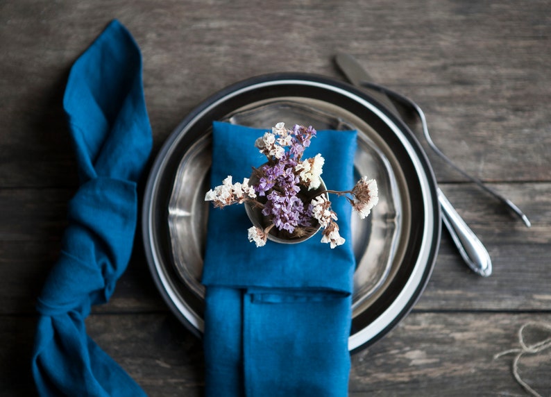 Peut inclure: Une serviette bleue est pli&eacute;e et plac&eacute;e sur une assiette en argent avec un bord noir. Un petit bouquet de fleurs s&eacute;ch&eacute;es est plac&eacute; sur la serviette. L'assiette est sur une table en bois.