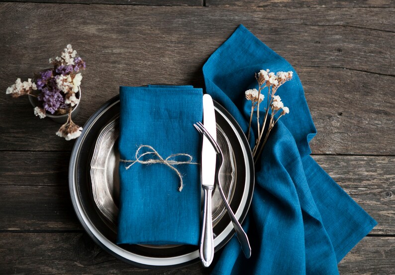 Peut inclure: Une serviette en lin bleu nou&eacute;e avec de la ficelle est plac&eacute;e sur une assiette argent&eacute;e et noire avec un couteau et une fourchette. L'assiette est pos&eacute;e sur une nappe en lin bleu.