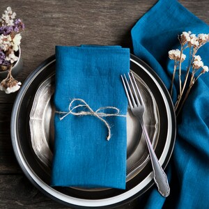 Peut inclure: Une serviette en lin bleu nou&eacute;e avec de la ficelle, plac&eacute;e sur une assiette argent&eacute;e et noire avec une fourchette. L'assiette est sur une table en bois avec un bouquet de fleurs s&eacute;ch&eacute;es.