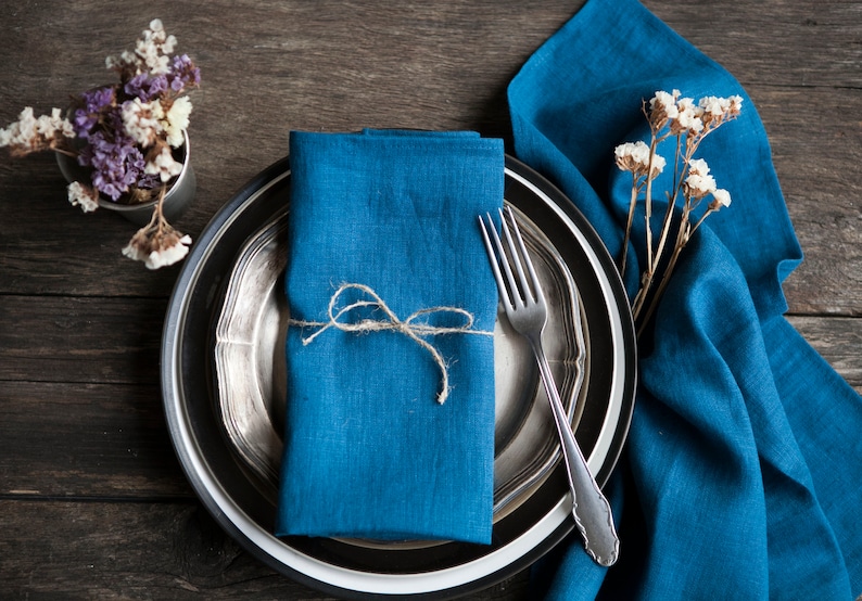 Peut inclure: Une serviette en lin bleu nou&eacute;e avec de la ficelle, plac&eacute;e sur une assiette argent&eacute;e et noire avec une fourchette. L'assiette est sur une table en bois avec un bouquet de fleurs s&eacute;ch&eacute;es.
