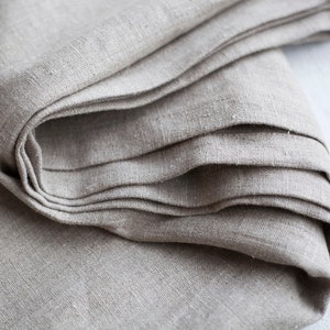 May include: A close-up of a folded piece of natural linen fabric. The fabric is a light beige color and has a slightly textured surface.