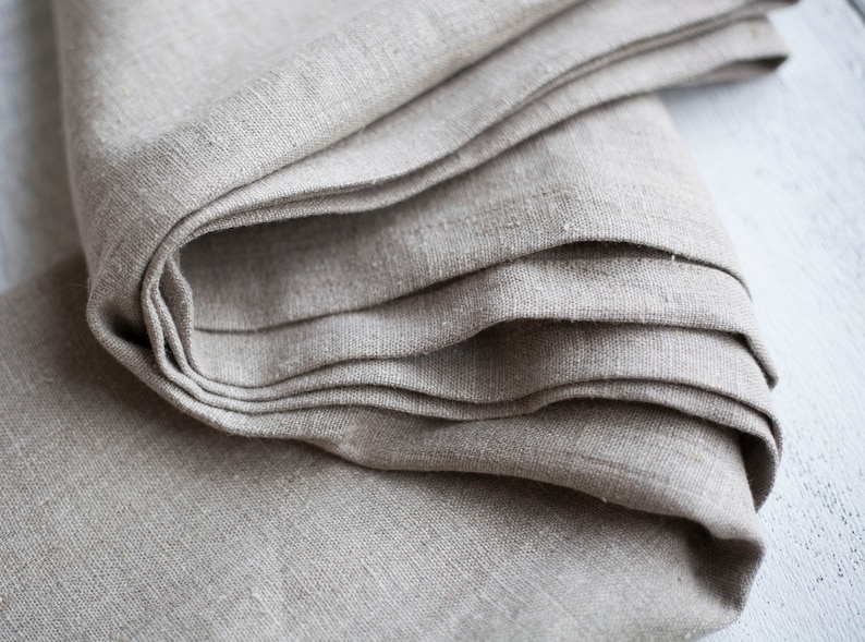 May include: A close-up of a folded piece of natural linen fabric. The fabric is a light beige color and has a slightly textured surface.