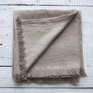 May include: A beige linen throw blanket with a fringed edge. The blanket is folded in half and laid flat on a white wooden surface.