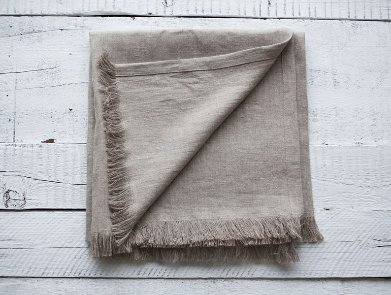 May include: A beige linen throw blanket with a fringed edge. The blanket is folded in half and laid flat on a white wooden surface.