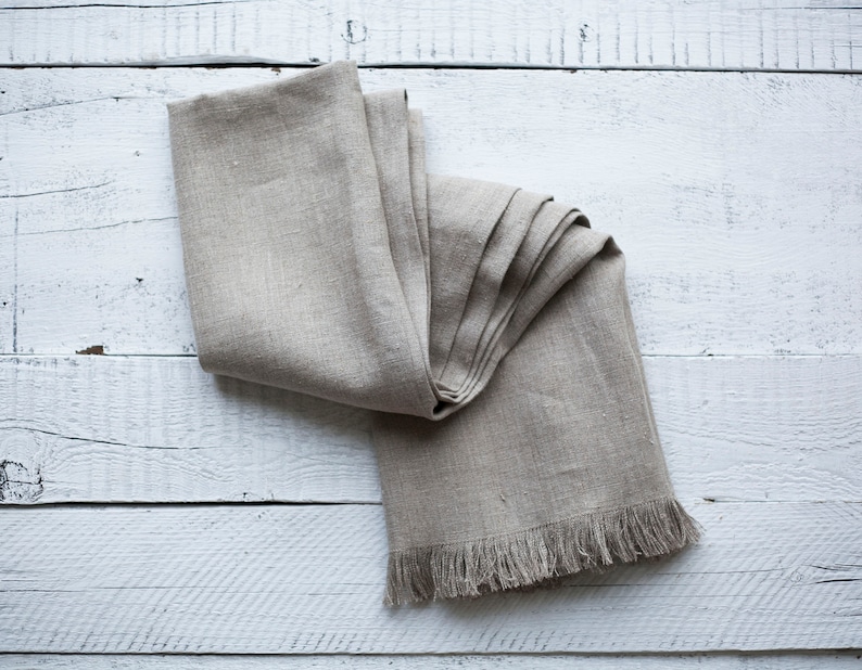May include: A folded linen table runner with a fringed edge, displayed on a white wooden surface.